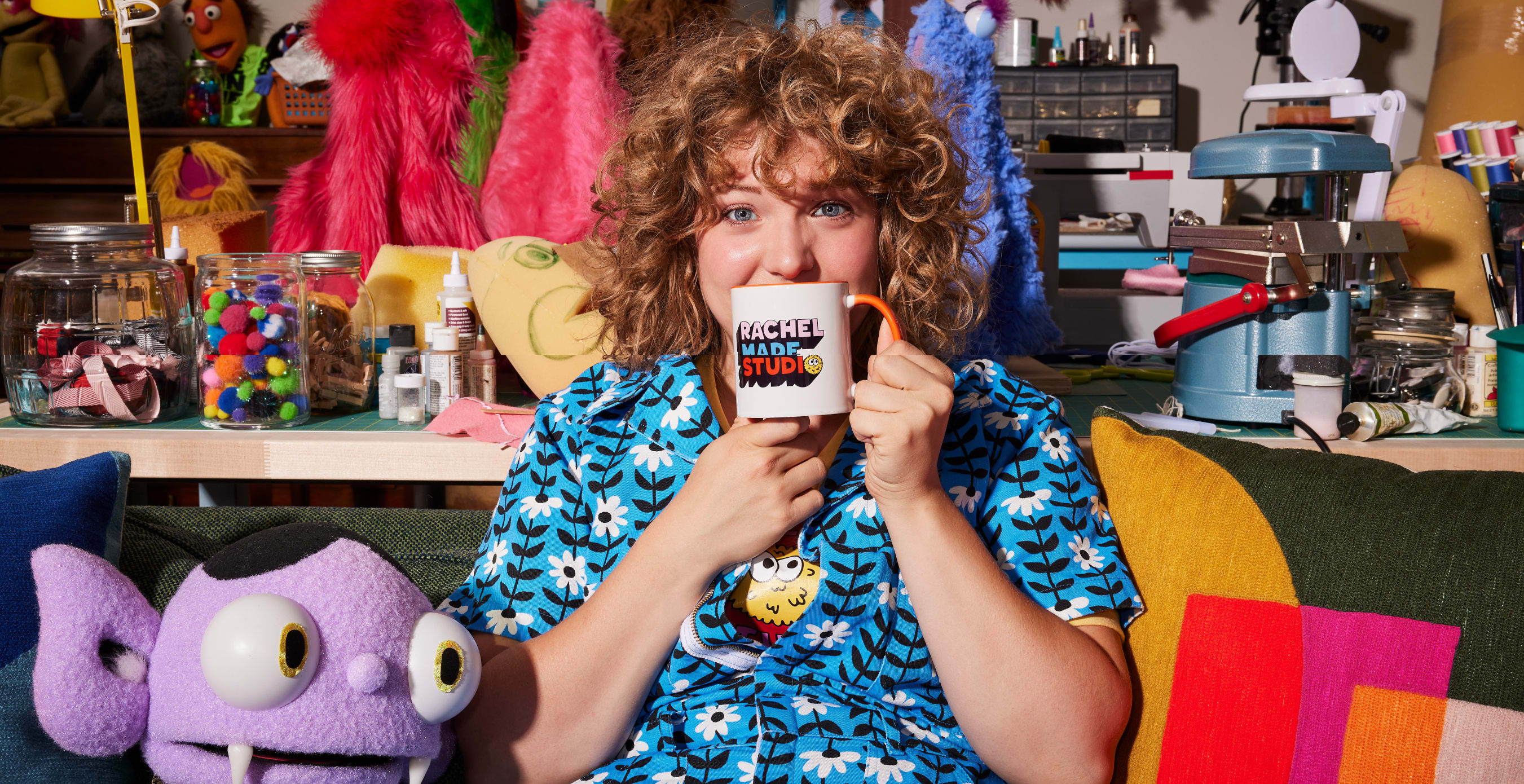 Business owner Rachel is sitting on a couch with her purple puppet next to her, holding a branded 'Rachel Made Studio' mug up to her mouth